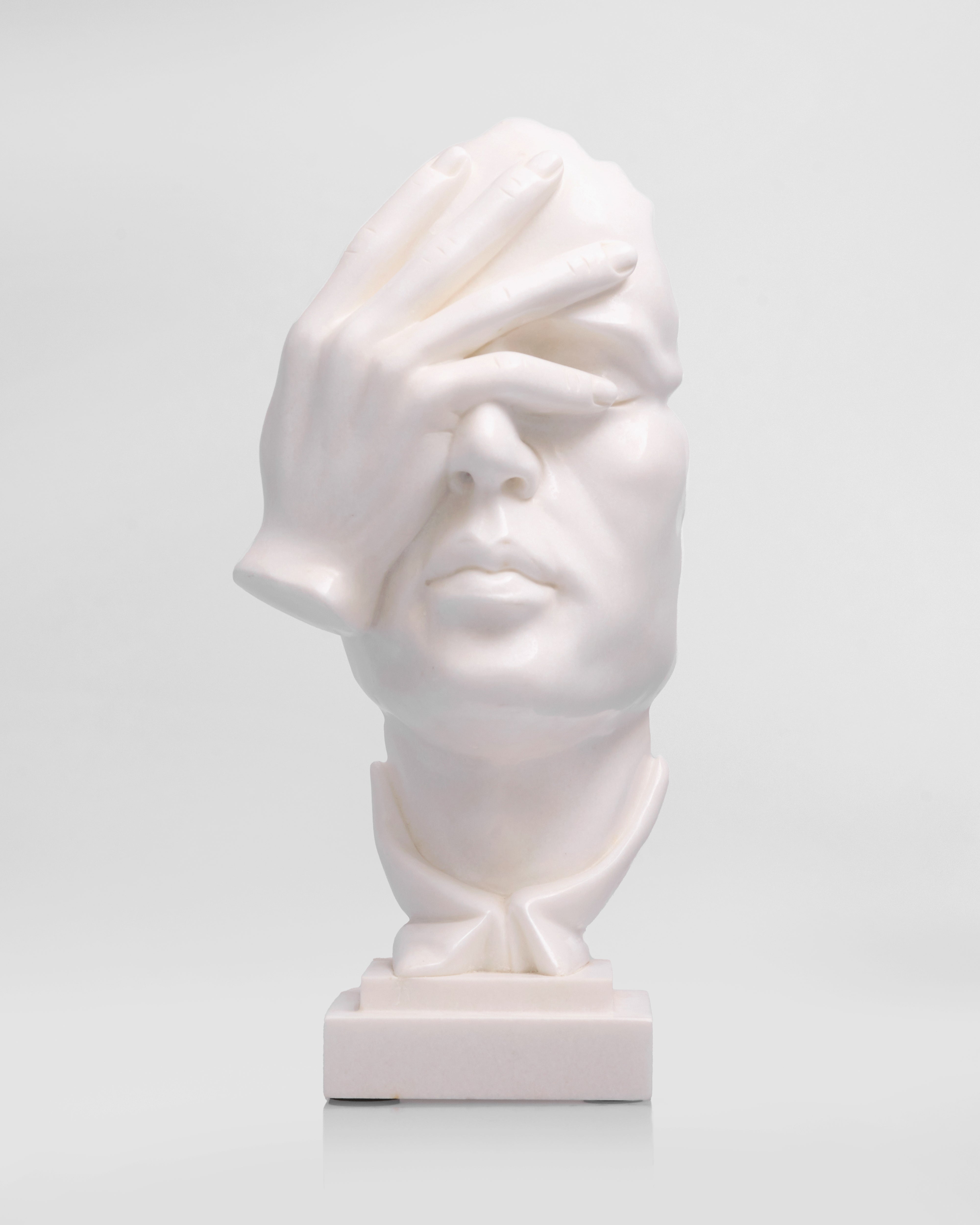 White ceramic sculpture of a face with one hand covering one eye on a light gray background