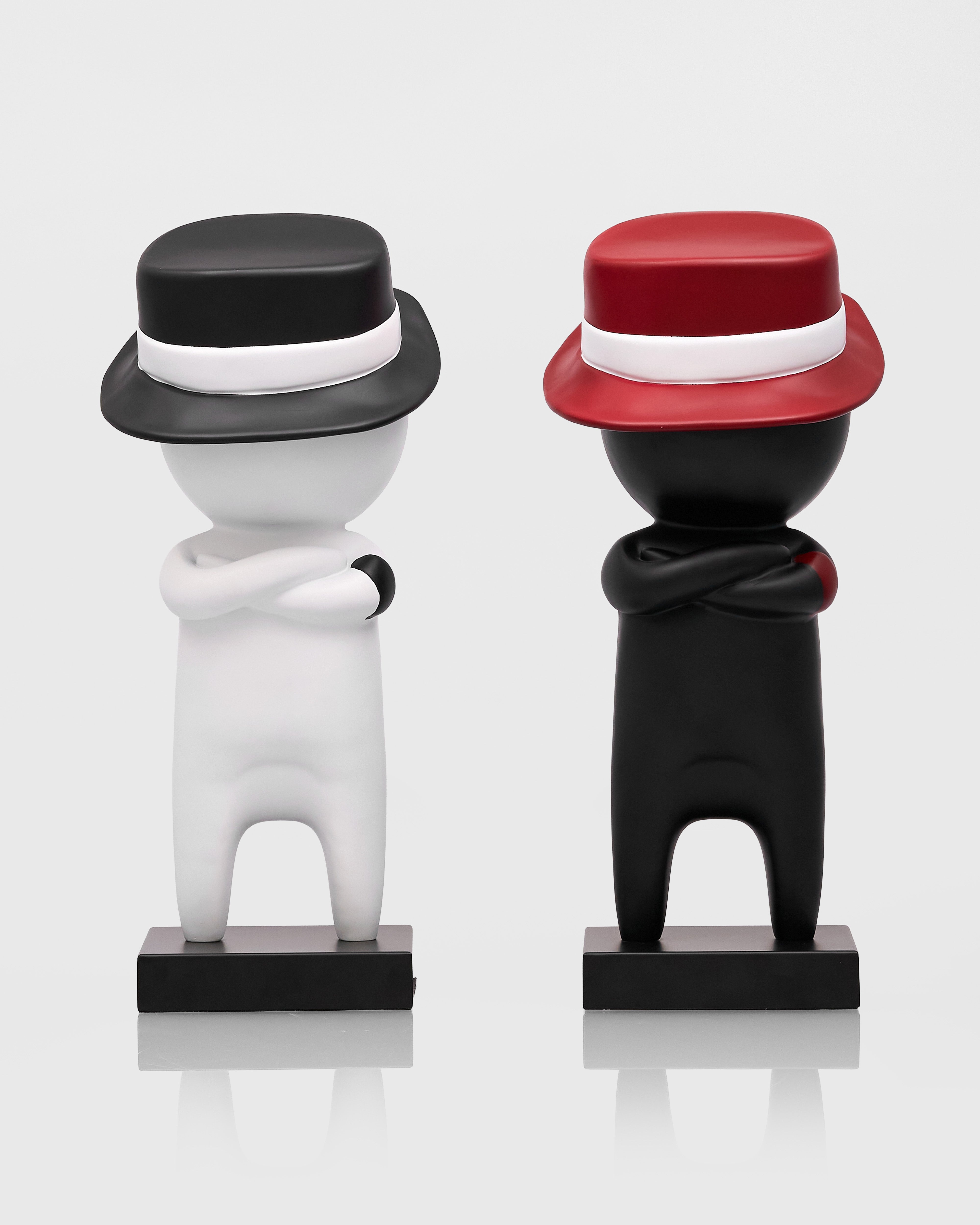 Two figurines, one white and one black, wearing hats on a reflective surface with a light gray background.