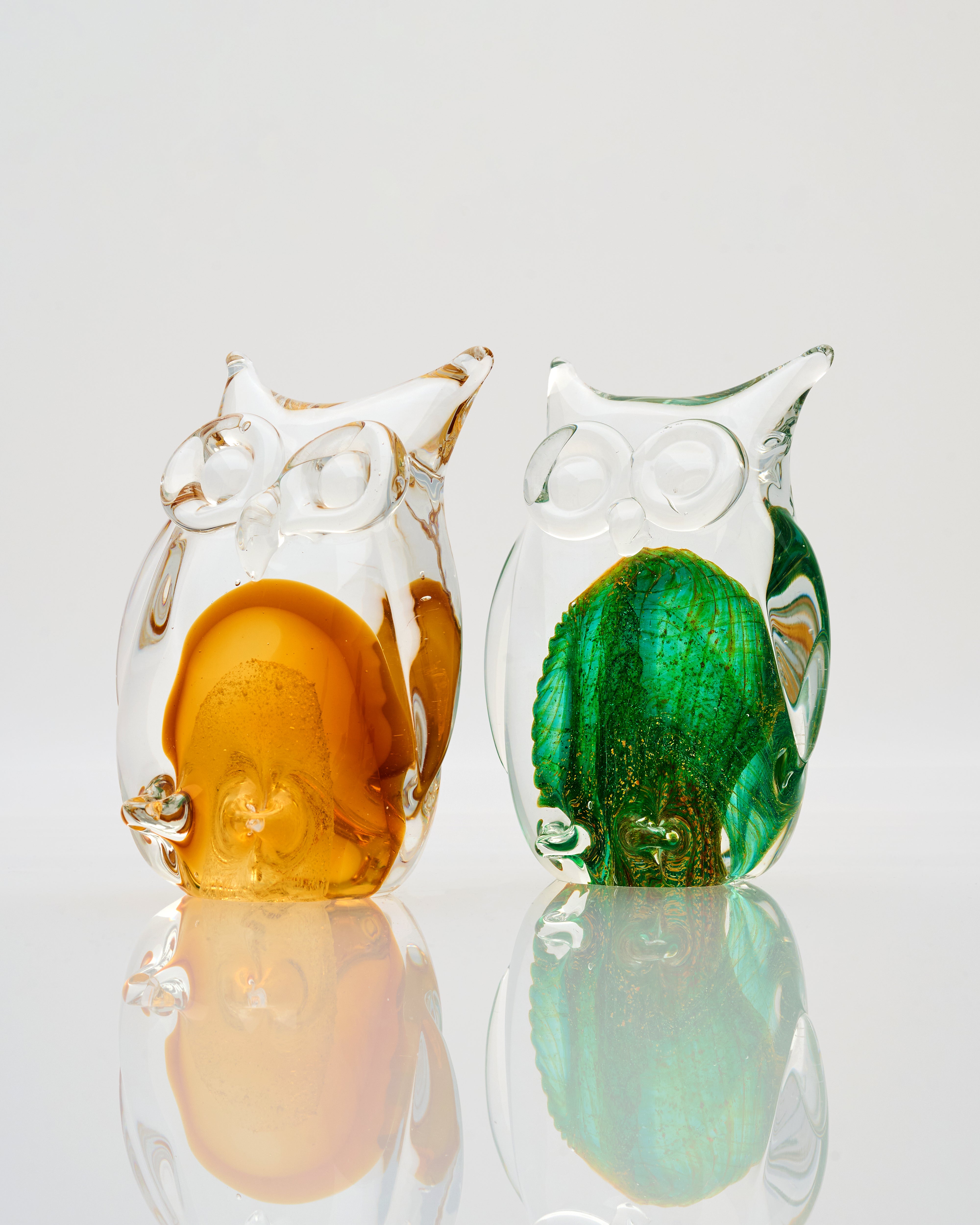 Two glass owl figurines, one orange and one green, on a reflective surface with a light gray background.