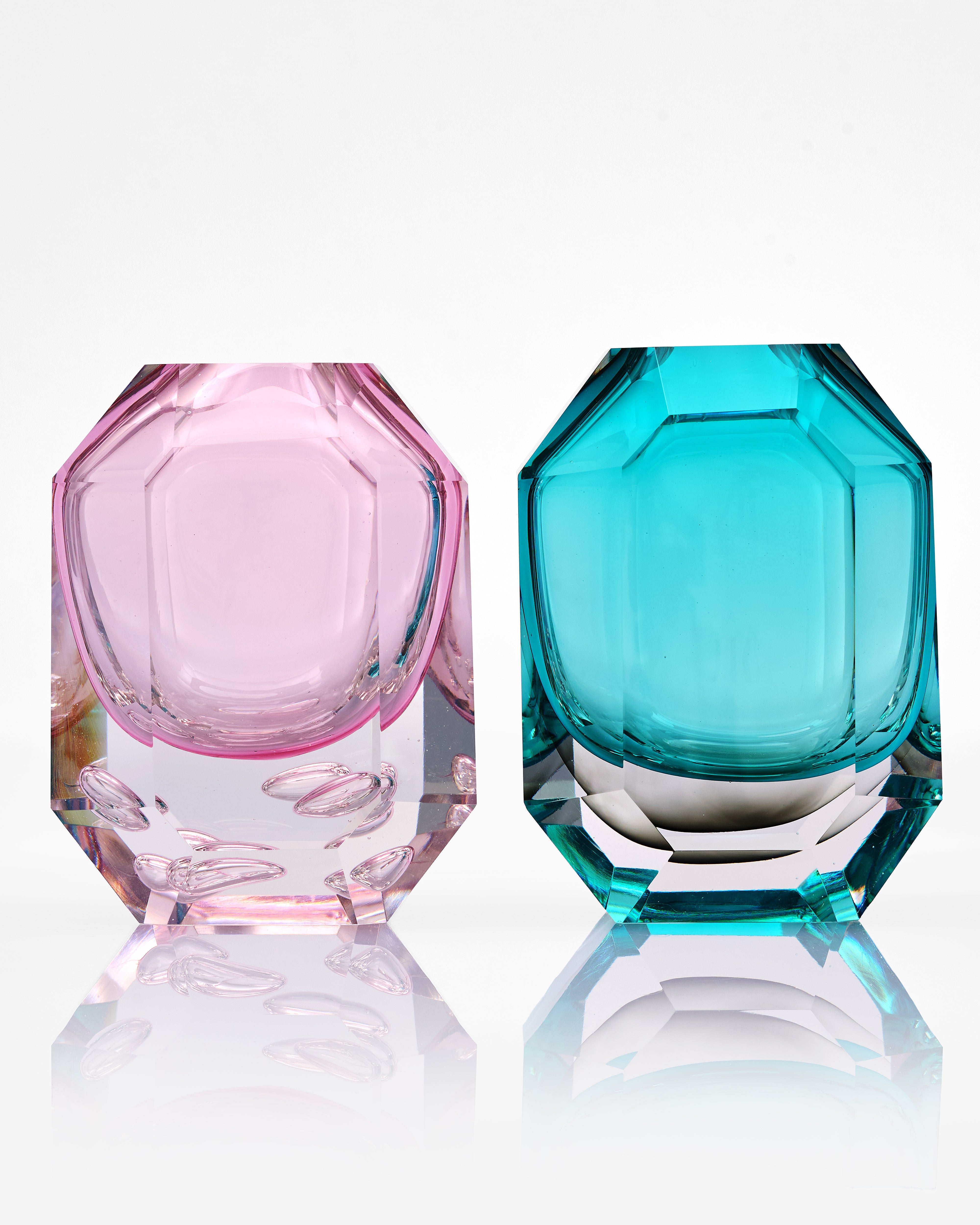 Two faceted glass containers, one pink and one blue, on a reflective surface with a white background.