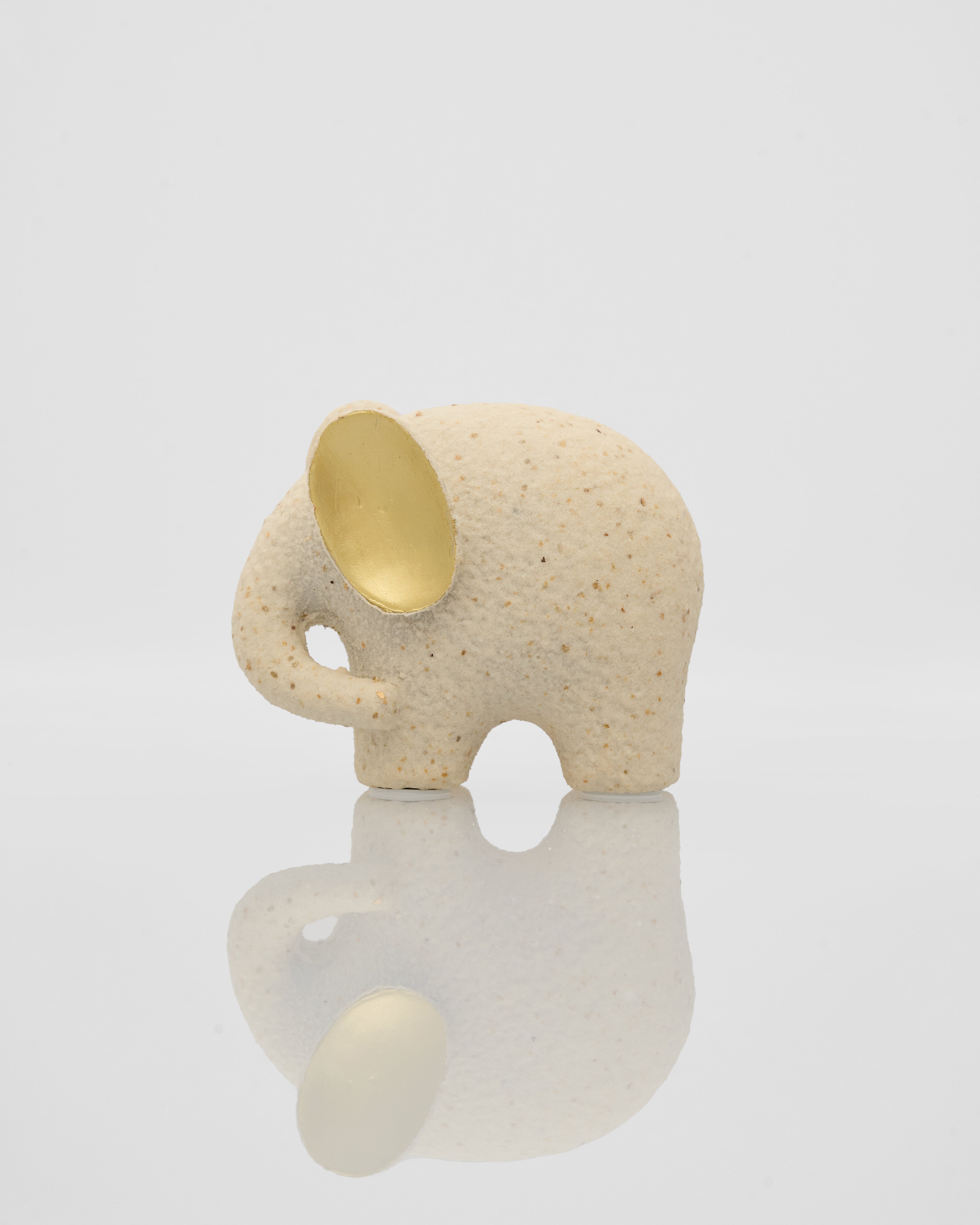 Small ceramic elephant figurine with a gold ear on a reflective surface.