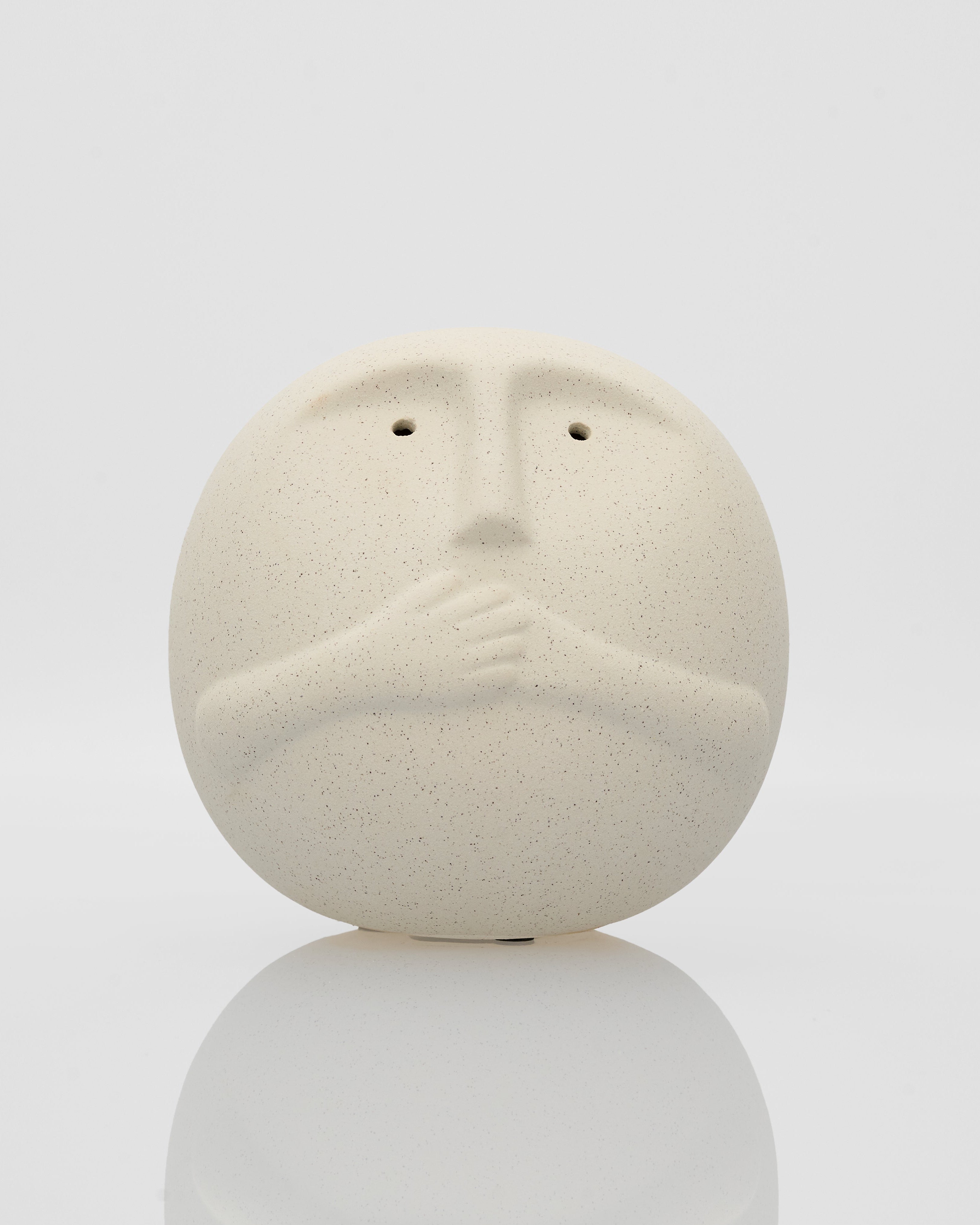 Ceramic face sculpture on a reflective surface with a white background