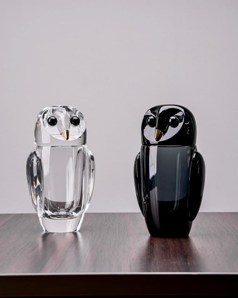 Two owl-shaped glass sculptures, one clear and one black, on a wooden surface with a gray background.