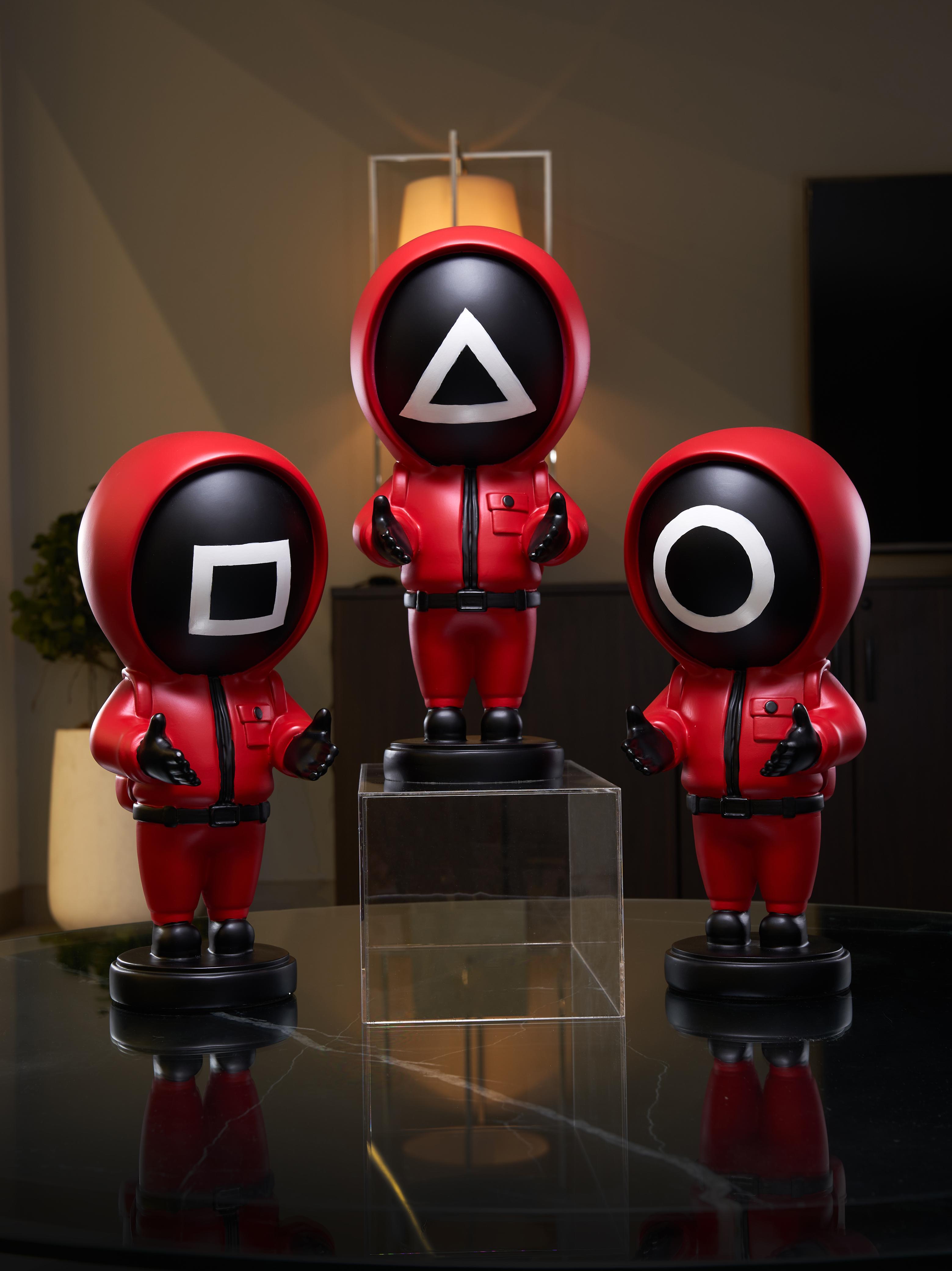 Three red and black character figurines of squid game series with different symbols on their faces, standing on a reflective surface