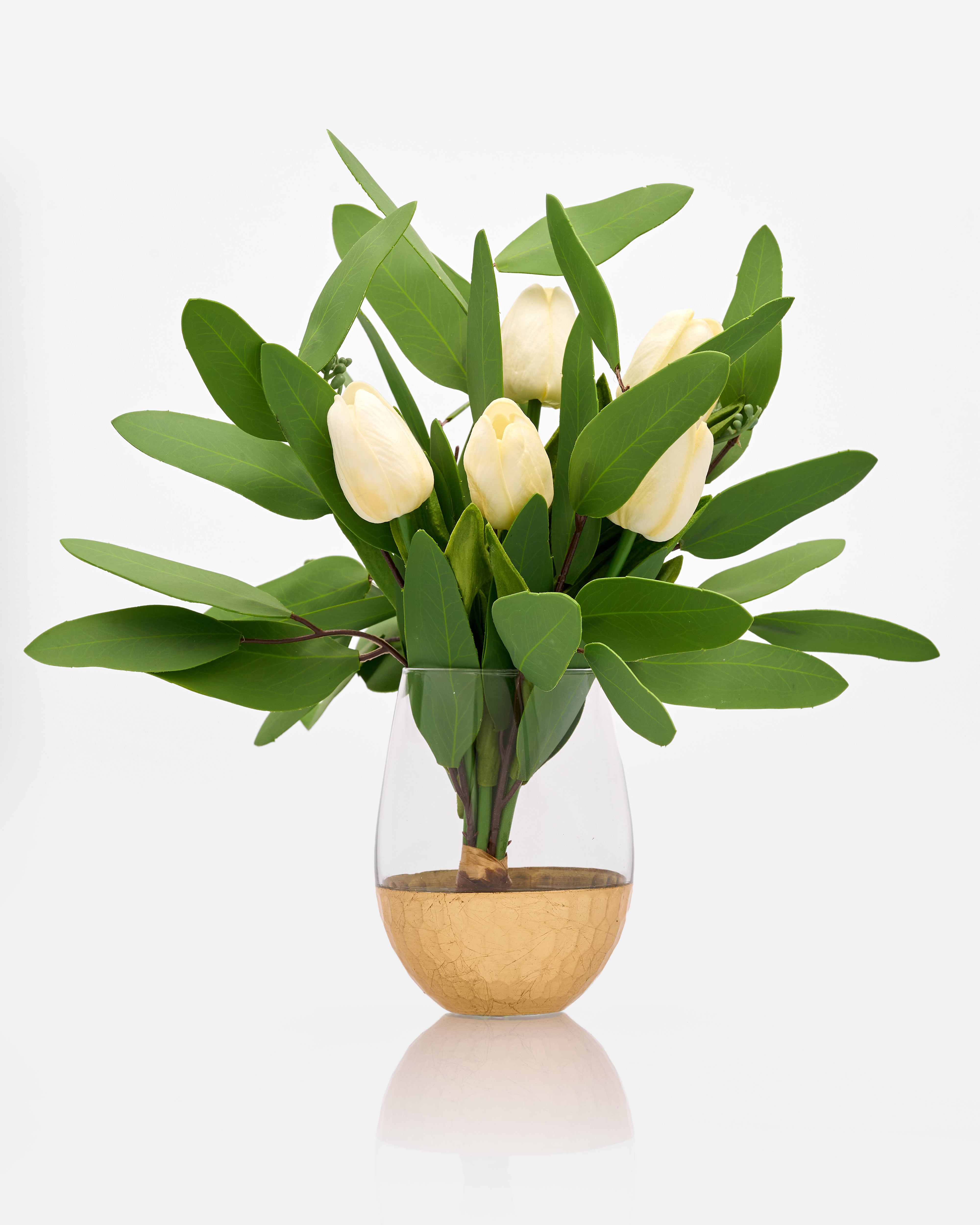 Clear vase with gold base containing white tulips and green leaves on a white background