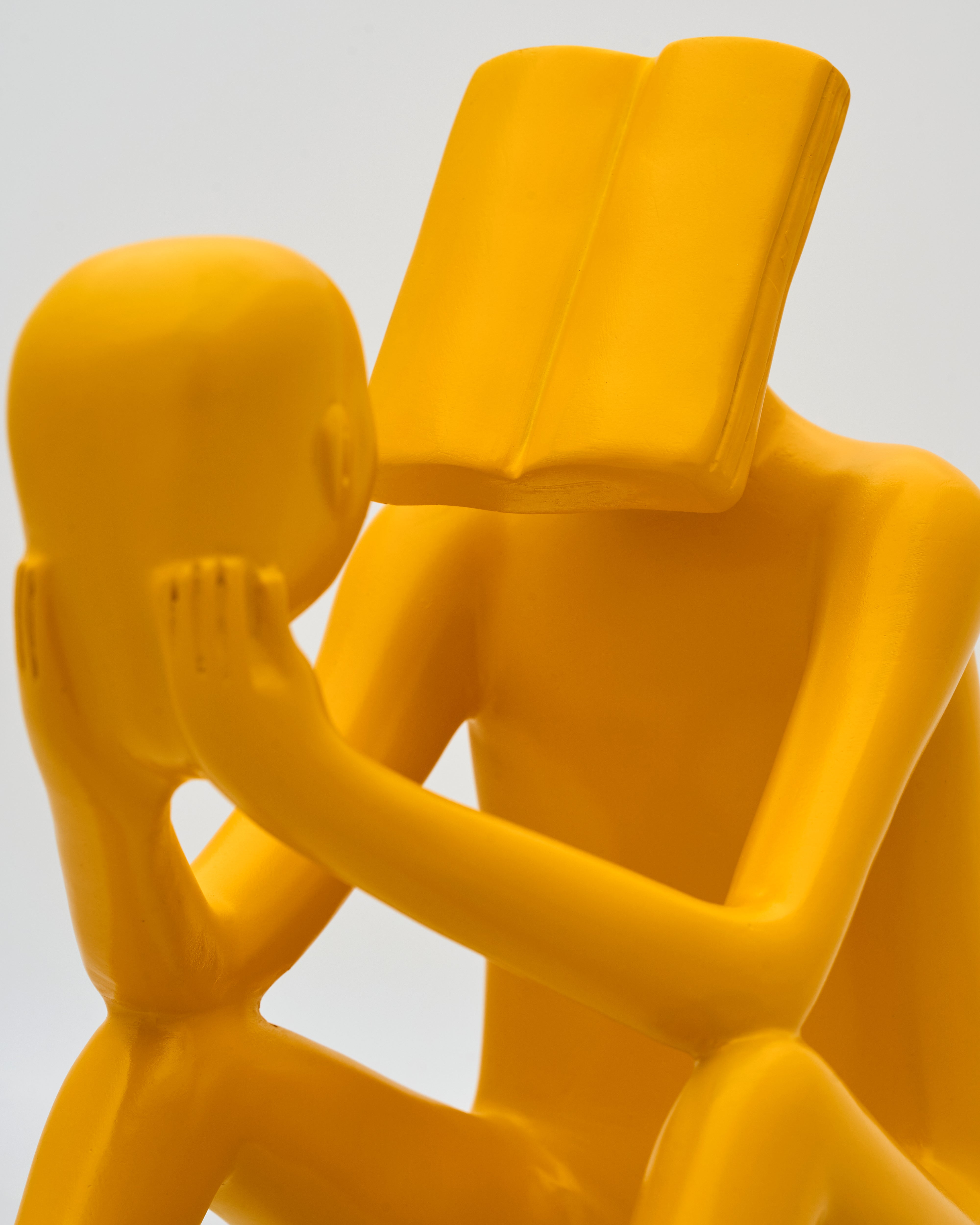 Yellow sculpture of two abstract figures in a close embrace with one figure holding a book. 