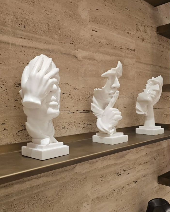 Three white abstract sculptures on a wooden shelf against a stone wall.