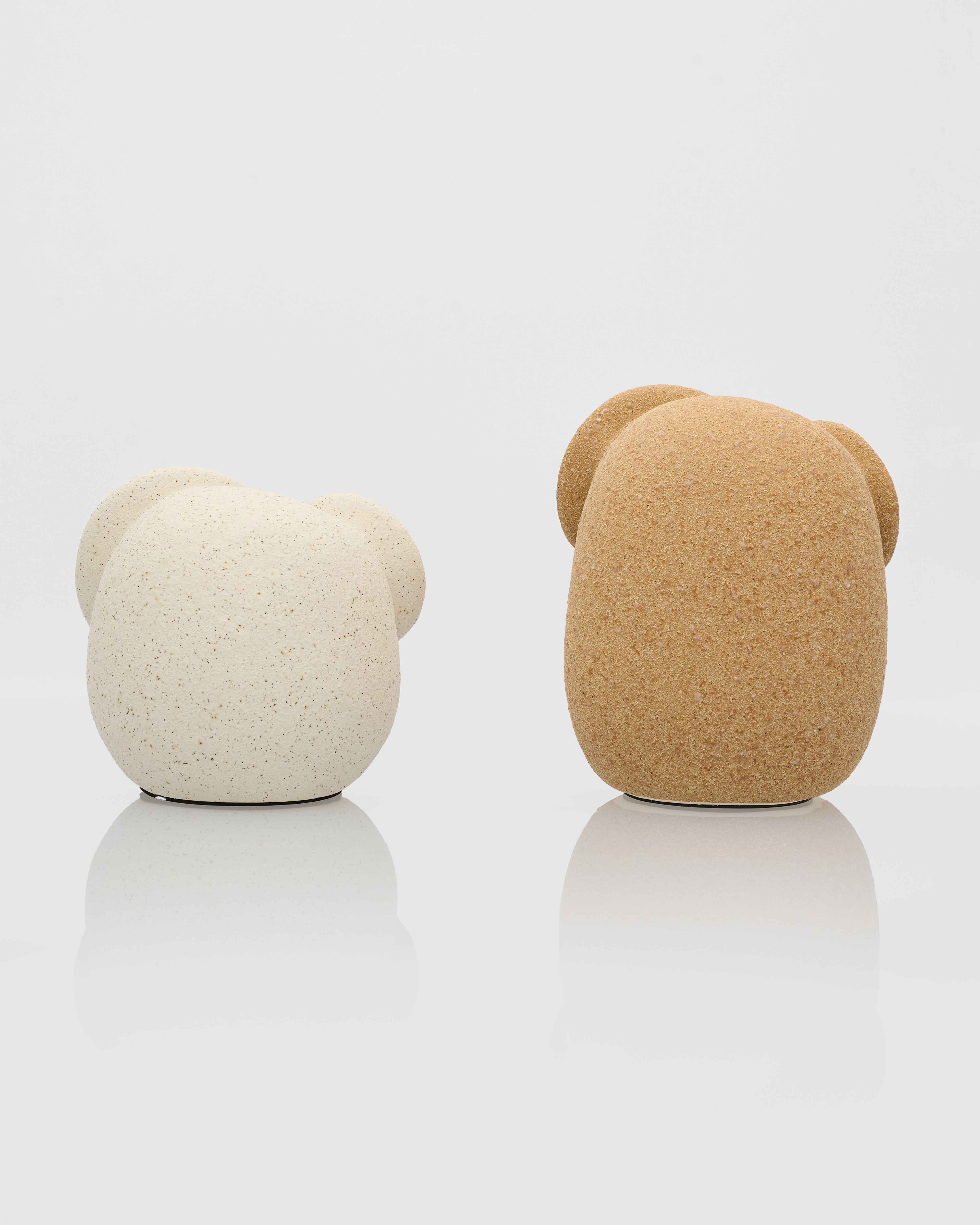Two bear-shaped objects, one beige and one white, on a reflective surface with a light gray background.